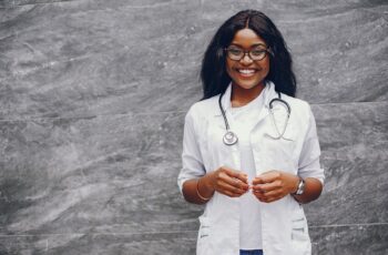 Stylish black doctor in a white uniform. Lady with stethoscope. Woman standing near gray wall